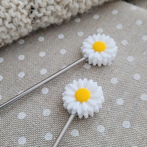 Op de afbeelding: Twee zilveren roerlepels met witte, madeliefjesvormige toppen en gele centra. De lepels liggen op een lichtbruine stoffen achtergrond met witte stippen.