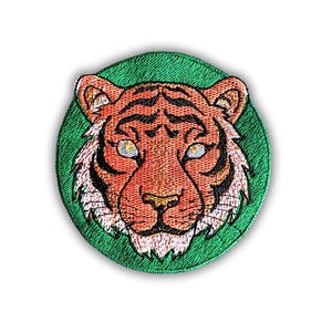 Puede incluir: Parche bordado con la cara de un tigre. El tigre tiene rayas naranjas y negras, detalles blancos y ojos amarillos. El parche está sobre un fondo verde circular, creando un contraste llamativo.
