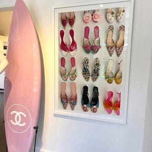 Może przedstawiać: Różowa deska surfingowa z białym logo Chanel stoi obok oprawionej ekspozycji różnych butów na wysokim obcasie. Buty są w różnych kolorach i stylach, w tym z kwiatowymi i wzorzystymi wzorami.