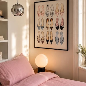 May include: A framed wall art print featuring a grid of fifteen illustrated shoes in various colors and styles, including pointed-toe flats, heels, and mules in shades of pink, black, leopard print, and blue, displayed in a black frame.