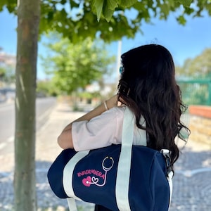 Può includere: Borsa da viaggio blu navy con cinghie bianche e la scritta "ENFERMERA" e un grafico di stetoscopio. La borsa è portata a tracolla. Lo sfondo comprende un albero e un marciapiede.