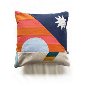 May include: Square cushion with a geometric design. The cushion features a navy blue triangle with a white star, orange and coral stripes, a light blue semi-circle, and a beige and teal base. The cushion is on a white background.