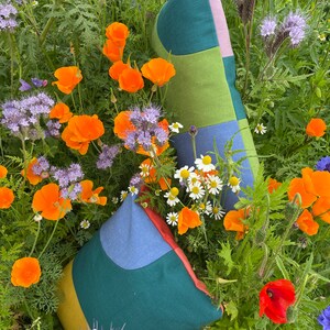 Peut inclure: Deux housses de coussin en patchwork color&eacute; avec des motifs g&eacute;om&eacute;triques dans des tons de vert, bleu, jaune et rose. Les coussins sont pos&eacute;s sur un lit de fleurs sauvages, dont des coquelicots orange et des fleurs bleues.