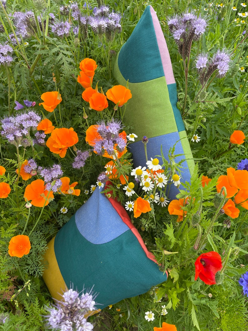 Peut inclure: Deux housses de coussin en patchwork color&eacute; avec des motifs g&eacute;om&eacute;triques dans des tons de vert, bleu, jaune et rose. Les coussins sont pos&eacute;s sur un lit de fleurs sauvages, dont des coquelicots orange et des fleurs bleues.