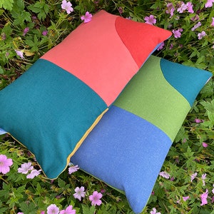 Peut inclure: Deux coussins rectangulaires avec des motifs g&eacute;om&eacute;triques dans des tons de bleu sarcelle, vert, bleu, jaune et corail. Les coussins sont pos&eacute;s sur un lit de verdure avec des fleurs roses.