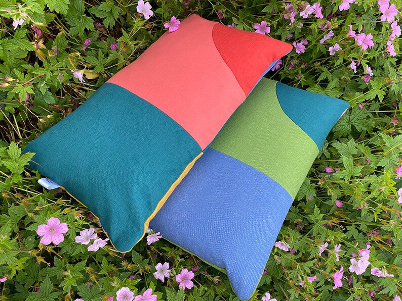 Peut inclure: Deux coussins rectangulaires avec des motifs g&eacute;om&eacute;triques dans des tons de bleu sarcelle, vert, bleu, jaune et corail. Les coussins sont pos&eacute;s sur un lit de verdure avec des fleurs roses.