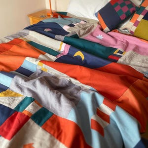 May include: A patchwork quilt featuring a vibrant geometric design with blocks of orange, navy blue, teal, light blue, pink, and beige. The fabric includes appliqued shapes such as a yellow crescent moon, a white star, and an orange arrow.