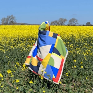 May include: A colorful geometric patterned quilt in shades of blue, yellow, red, green, and white. The quilt is draped over a person standing in a field of yellow flowers.