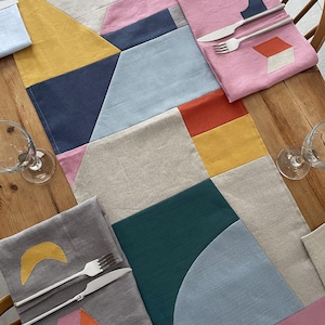 Puede incluir: Un juego de mesa geométrico con un camino de mesa de lino beige y servilletas a juego. Las servilletas están adornadas con formas coloridas e incluyen cubiertos. La paleta de colores incluye rosa, amarillo, azul y verde.