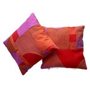 Puede incluir: Dos cojines cuadrados con un diseño de patchwork de varias texturas de tela en tonos rojos, naranjas y morados. Los cojines están construidos con múltiples formas geométricas cosidas entre sí, creando una superficie vibrante y multicolor.