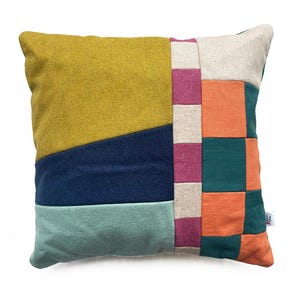 May include: Square cushion with a patchwork design. The cushion features sections of mustard yellow, navy blue, teal, orange, and green fabric. A vertical stripe of pink and white squares runs down the centre. The cushion is square and made of a textured fabric.