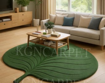 May include: A green leaf-shaped rug with detailed vein patterns dominates a living room scene. The rug is placed on a wooden floor, with a wooden coffee table, a beige sofa, and a wooden cabinet in the background. The text "KGNCARPET" is visible on the rug.