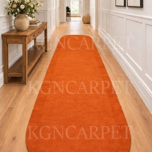 May include: An orange runner rug in a hallway. The rug is a solid color and has rounded ends. The hallway has light wood flooring, white walls, and a wooden console table with a mirror.