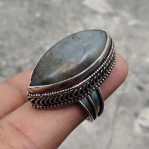 May include: A silver ring featuring a large, oval-shaped, iridescent labradorite stone. The stone is set in a detailed, ornate silver band with a textured design. The ring is a statement piece of jewelry.