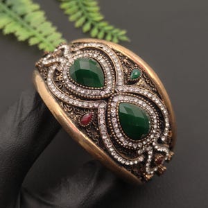 May include: A wide cuff bracelet in an antique bronze tone featuring two prominent teardrop-shaped dark green stones. The surface is intricately detailed with small clear crystals and accented by tiny red stones, creating a textured, ornate appearance.