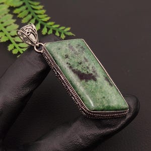 May include: A rectangular pendant featuring a polished green and black stone set in a decorative silver-toned metal frame with a filigree bail. The stone has a mottled texture with dark inclusions throughout the surface of the gemstone.