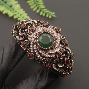 May include: An ornate hinged cuff bracelet in an antique gold-tone finish, featuring a large central emerald green faceted stone surrounded by swirling filigree patterns, clear white crystals, and smaller red and green accent stones.