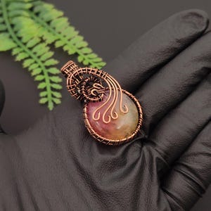 Peut inclure: Un pendentif composé d'une pierre ronde aux nuances de rose, jaune et orange, entourée d'un fil de cuivre finement travaillé. Le design en fil métallique comprend un cadre circulaire avec des motifs en spirale s'étendant vers le haut.