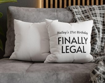 May include: Two white square cushions on a grey sofa. One cushion has the text "Hailey's 21st Birthday FINALLY LEGAL" in black. The other cushion is plain white. A grey blanket with a black and white plaid pattern is draped over the sofa.