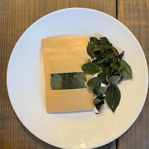 Puede incluir: Un plato blanco sobre una superficie de madera. Una pequeña bolsa de papel marrón con una ventana transparente, llena de hojas verdes. Un pequeño montón de hojas verdes está al lado de la bolsa.