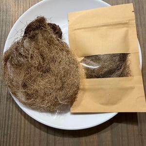 Könnte beinhalten: Auf einem weißen Teller befindet sich eine Masse aus braunem, faserigem Material, möglicherweise Barthaar. Daneben liegt ein kleiner brauner Papierbeutel mit einem transparenten Fenster, der ebenfalls das gleiche Material enthält. Die Gegenstände sind vor einem Holzgrund platziert.