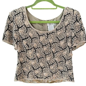 Puede incluir: Top beige de manga corta con estampado floral. El top presenta un escote redondo y est&aacute; adornado con bordados florales negros y beige. La prenda est&aacute; colgada de una percha verde sobre un fondo blanco.