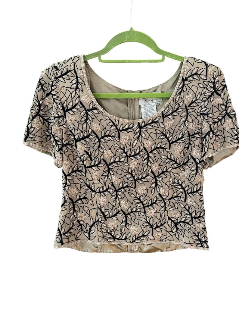 Puede incluir: Top beige de manga corta con estampado floral. El top presenta un escote redondo y est&aacute; adornado con bordados florales negros y beige. La prenda est&aacute; colgada de una percha verde sobre un fondo blanco.
