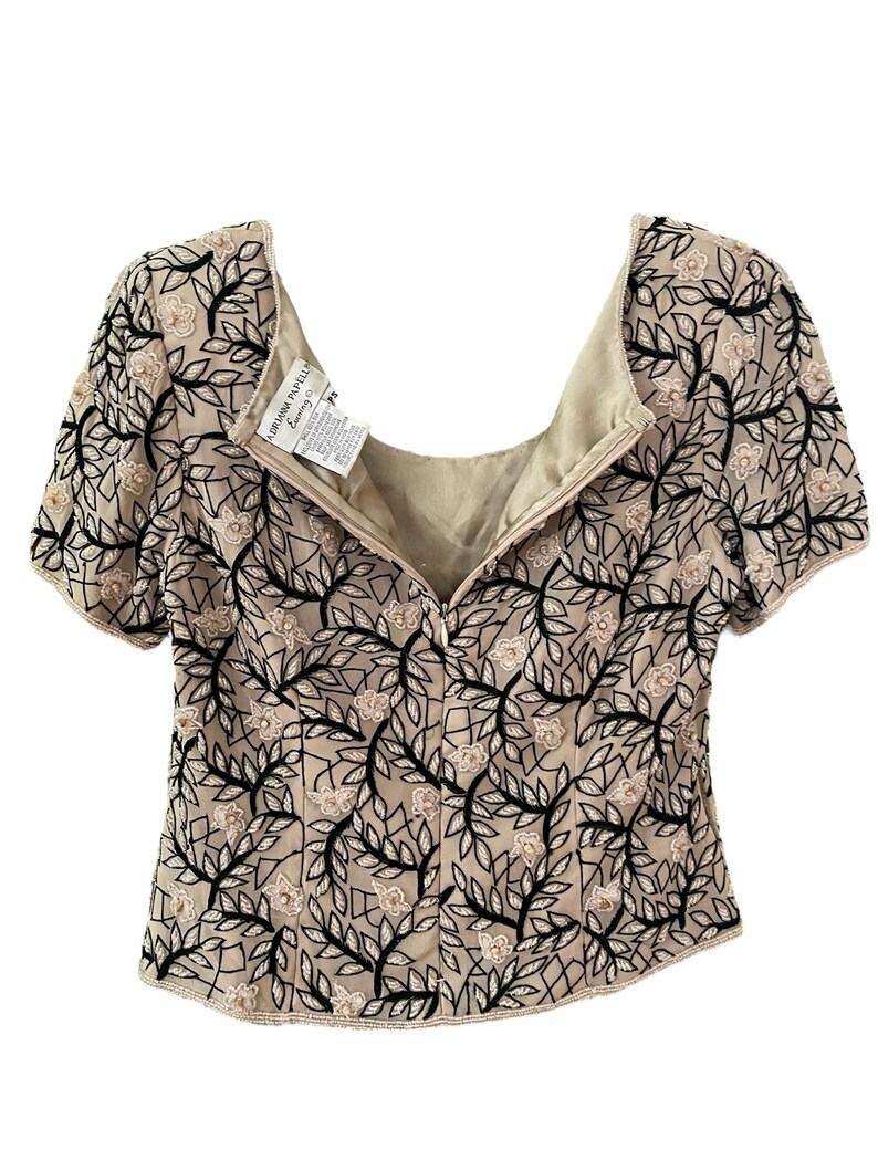 Puede incluir: Top beige de manga corta con cuello cuadrado. El top est&aacute; adornado con un dise&ntilde;o floral en blanco y negro. La etiqueta dice "Adrianna Papell". El top tiene cierre de cremallera en la espalda.