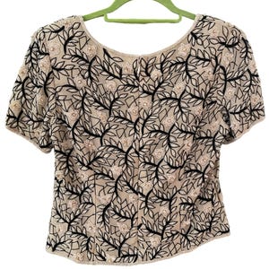 Puede incluir: Top beige de manga corta con estampado floral. El top presenta un delicado dise&ntilde;o de vides y hojas negras, con peque&ntilde;as flores de color claro. El escote y las mangas est&aacute;n ribeteados con peque&ntilde;as cuentas. El top est&aacute; colgado de una percha verde.