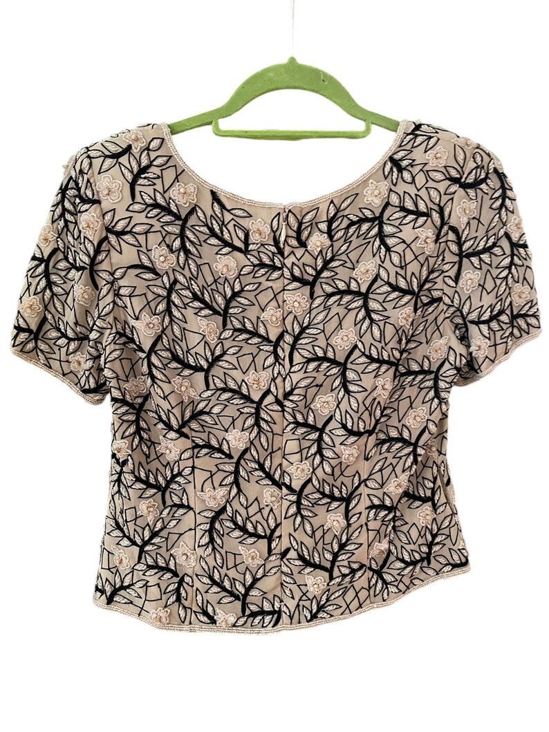Puede incluir: Top beige de manga corta con estampado floral. El top presenta un delicado dise&ntilde;o de vides y hojas negras, con peque&ntilde;as flores de color claro. El escote y las mangas est&aacute;n ribeteados con peque&ntilde;as cuentas. El top est&aacute; colgado de una percha verde.