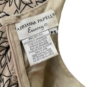 Puede incluir: Primer plano de la etiqueta de un vestido Adrianna Papell Evening. La etiqueta blanca tiene texto negro, incluyendo el nombre de la marca y "Evening". El vestido es beige con bordados florales negros y blancos. La etiqueta indica que el vestido est&aacute; hecho en India.