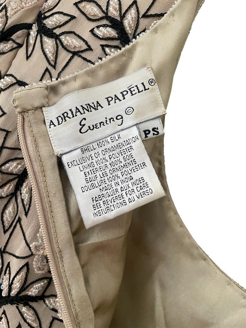 Puede incluir: Primer plano de la etiqueta de un vestido Adrianna Papell Evening. La etiqueta blanca tiene texto negro, incluyendo el nombre de la marca y "Evening". El vestido es beige con bordados florales negros y blancos. La etiqueta indica que el vestido est&aacute; hecho en India.