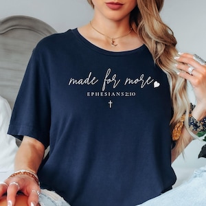 May include: Navy blue t-shirt with the text "made for more" in a handwritten style, along with the Bible verse Ephesians 2:10 and a small cross. The shirt is a comfortable, casual style.