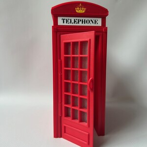 May include: A red miniature telephone booth with an open door. The booth has a crown emblem and the word "TELEPHONE" in white. The door features a grid of rectangular panes.
