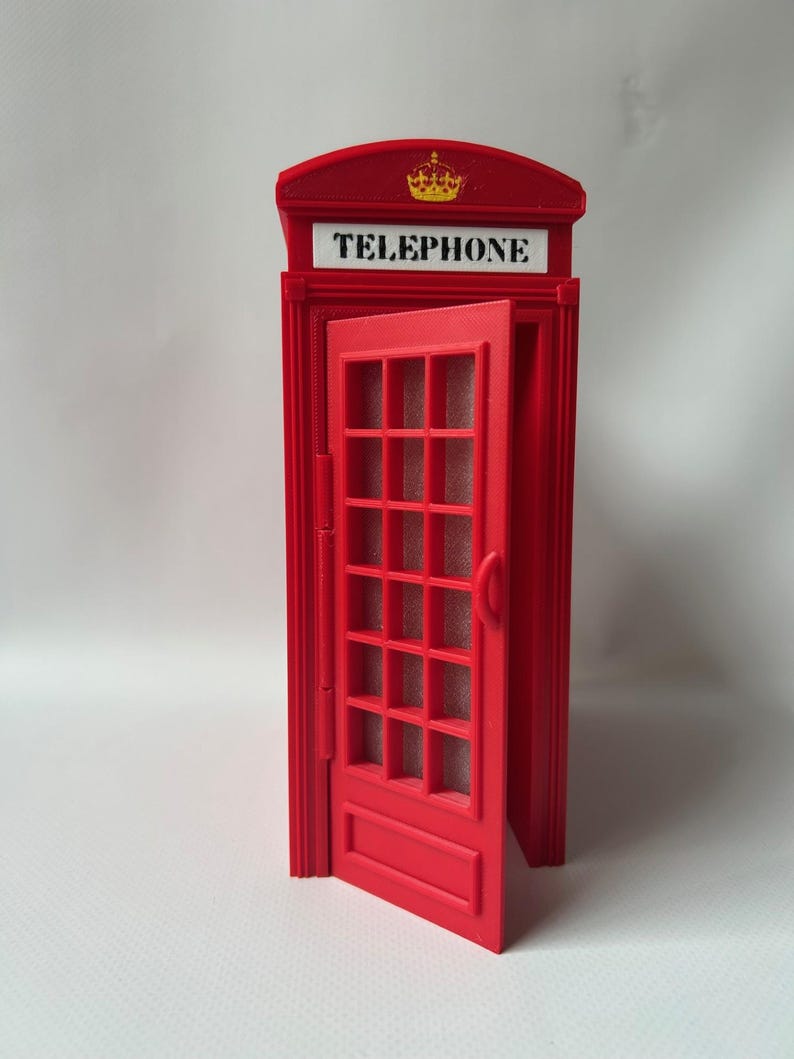May include: A red miniature telephone booth with an open door. The booth has a crown emblem and the word "TELEPHONE" in white. The door features a grid of rectangular panes.