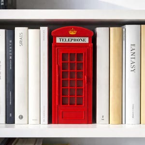 May include: A red miniature telephone booth stands on a white shelf, surrounded by books of various colors and sizes. The booth has a crown on top and the word "TELEPHONE" is displayed. The books are arranged neatly.