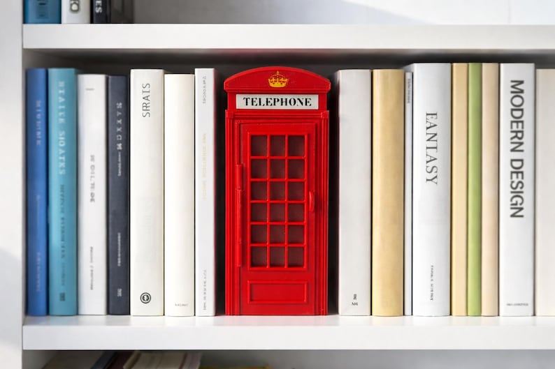 May include: A red miniature telephone booth stands on a white shelf, surrounded by books of various colors and sizes. The booth has a crown on top and the word "TELEPHONE" is displayed. The books are arranged neatly.