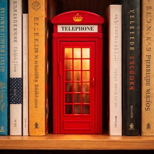 May include: A miniature red telephone booth with the word "TELEPHONE" on top, illuminated from within, sits on a wooden shelf among various books. The booth has a crown emblem above the text.