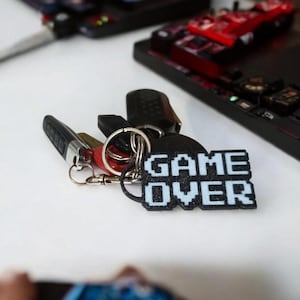 May include: A set of keys with a "GAME OVER" keychain. The keychain is black with white pixelated text. Other keys and key fobs are attached to the keyring. In the background are black gaming consoles and a red toy car.
