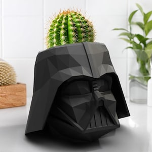 May include: A black, geometric Darth Vader helmet-shaped planter holding a green cactus with yellow spines. The planter is made of a matte material and sits on a white surface. The cactus is round and spiky, contrasting with the smooth, angular planter.