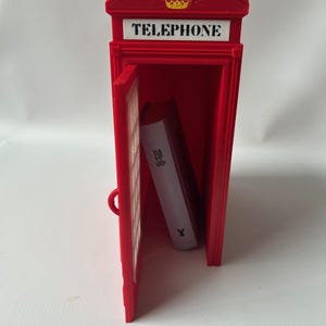 May include: A red miniature telephone booth with the word "TELEPHONE" on top and a gold crown. The door is open, revealing a white book inside. The book has the text "HA ME VAP" on the side.