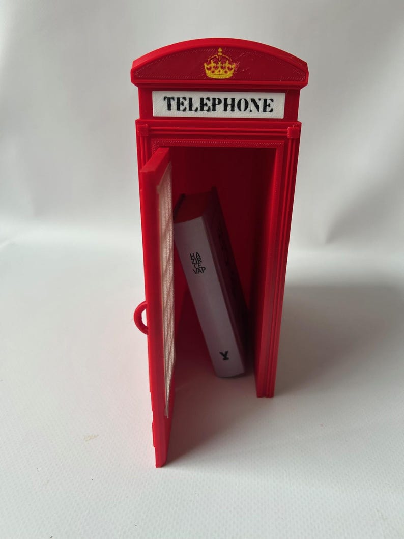 May include: A red miniature telephone booth with the word "TELEPHONE" on top and a gold crown. The door is open, revealing a white book inside. The book has the text "HA ME VAP" on the side.
