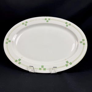 May include: White oval serving platter with a green and light green geometric design around the rim. The platter is displayed on a clear plastic stand against a black background. A classic piece for serving.