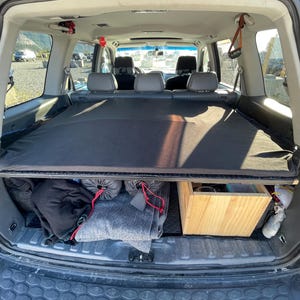 Puede incluir: La parte trasera abierta de un SUV negro, que revela un área de carga con una cubierta negra. En el interior, hay sacos de dormir enrollados, una manta gris y una caja de madera. El interior del vehículo incluye asientos grises y un techo corredizo.
