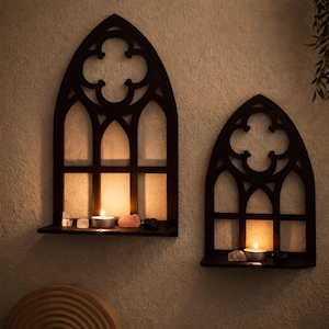 May include: Two dark brown wooden wall sconces in the shape of gothic windows. Each sconce has a small shelf holding a lit candle and crystals. The sconces are mounted on a textured beige wall, creating a warm, inviting ambiance.