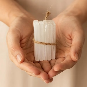 May include: A bundle of white selenite crystals, secured with natural twine. The crystals are held in open hands, highlighting their translucent, layered structure. The twine is wrapped around the middle and tied at the top.