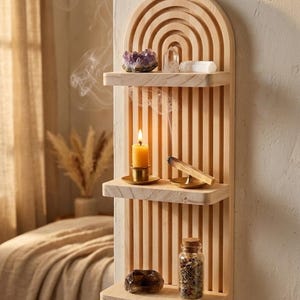 Puede incluir: Estanter&iacute;a de pared de madera con la parte superior arqueada y tres estantes. Los estantes contienen cristales, una vela encendida, un palito de humo y una peque&ntilde;a botella de vidrio con un corcho. La estanter&iacute;a es de madera clara.