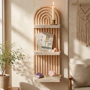 Puede incluir: Estanter&iacute;a de pared de madera clara con parte superior arqueada y tres estantes. Una vela encendida en el estante superior, cartas del tarot en el medio y cristales en el inferior.