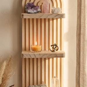 Puede incluir: Estanter&iacute;a de madera en forma de arco&iacute;ris con tres niveles, que muestra cristales, una vela encendida, un s&iacute;mbolo Om de bronce y un manojo de salvia. La estanter&iacute;a es de madera clara y est&aacute; montada en una pared beige.
