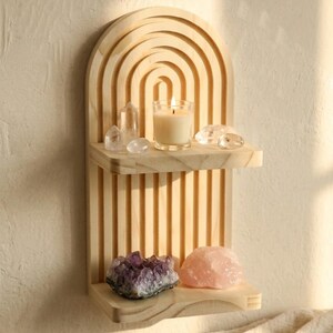 Puede incluir: Una estanter&iacute;a de pared de madera clara con una parte superior arqueada y dos estantes. El estante superior contiene una vela encendida en un vaso, cristales transparentes y un cristal de cuarzo. El estante inferior muestra una amatista p&uacute;rpura y un cristal de cuarzo rosa.